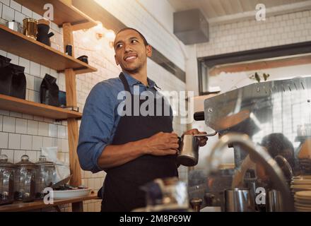 Gemischter Barista, der Milch für Kaffee aufschäumt. Bistro-Mitarbeiter, der Milch für eine Tasse Kaffee aufschäumt. Kaffeeeshop-Assistent hält eine Kanne, um Milch aufzuschäumen Stockfoto