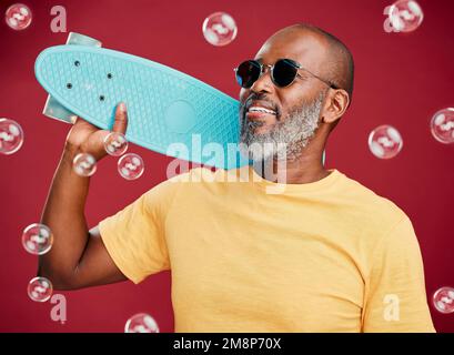 Ein reifer afroamerikanischer Mann steht mit einem Skateboard umgeben von Blasen vor einem roten Studiohintergrund. Ein glücklicher und sorgenfreier Schwarzer trägt ihn Stockfoto