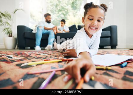 Ein kleines Mädchen malt mit Malstiften, die auf dem Boden des Wohnzimmers liegen, während ihr Vater und ihr Bruder sich auf dem Sofa entspannen. Ein kleines Mädchen, das sich dabei einfärbt Stockfoto