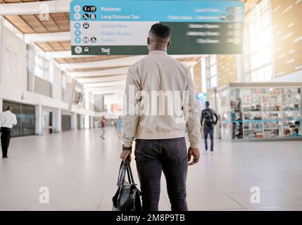 Rückansicht eines afroamerikanischen Geschäftsmanns, der allein reist und in einem Bahnhof steht, während er seine Reisezeiten auf dem Brett überprüft Stockfoto