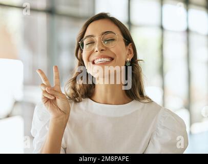Junger, aufgeregter Profi mit Brille, der Frieden seufzt. Verspielte Geschäftsfrau, Unternehmerin, die in einem Büro ein Siegesschild zeigt Stockfoto