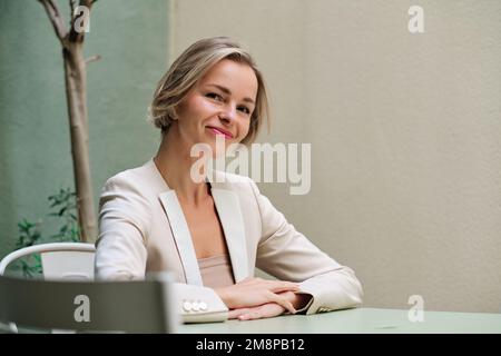 Eine Geschäftsfrau, die in die Kamera schaut und lächelt, während sie auf einem Tisch sitzt. Stockfoto