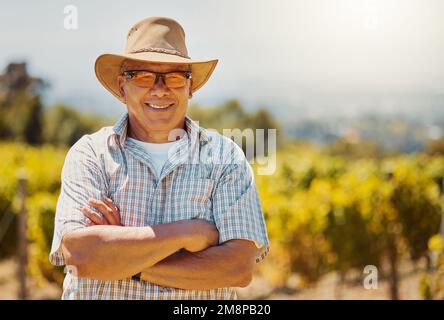 Ein selbstbewusster Bauer mit einem lächelnden Senior-Mixed-Rassen, der mit gekreuzten Armen auf seinem Weinberg steht. Hispanischer alter Mann mit seinen Armen Stockfoto