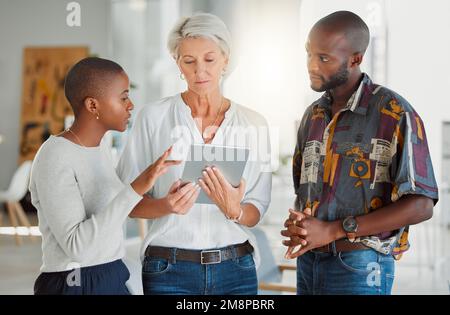 Drei fokussierte Geschäftsleute, die zusammen ein digitales Tablet bei der Arbeit verwenden. Reife weiße Geschäftsfrau spricht und zeigt ihr afroamerikanerin Stockfoto