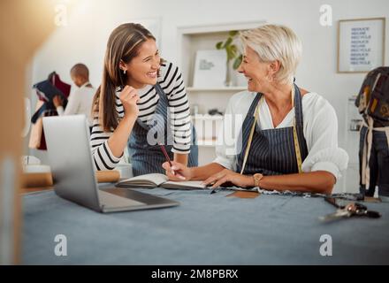 Zwei fröhliche Bekleidungsdesigner verwenden einen Laptop und schreiben in ein Notizbuch, während sie in einem Meeting mit Material arbeiten. Weiße und gemischte Rasse Stockfoto