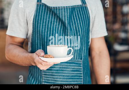 Nahaufnahme eines hispanischen Kellners, der eine Tasse Kaffee mit einem Keks serviert, während er in einem Café arbeitet. Die Hände des Baristas halten die Teetasse auf einer mit gefüllten Untertasse Stockfoto