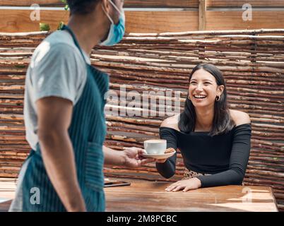 Glückliche junge hispanische Frau, die eine Tasse Kaffee von einem Kellner mit Gesichtsmaske in einem lokalen Café bekam. Gemischte Rasse, die sich darauf freut, warm serviert zu werden Stockfoto