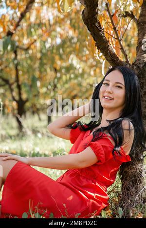 Gehen Sie im Garten spazieren. Eine brünette Frau in einem roten Kleid sitzt auf dem Gras, lehnt sich an einen Baumstamm und lächelt fröhlich. Stockfoto
