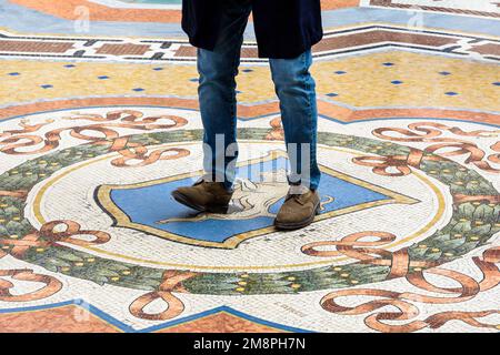 Ein Tourist, der das glückliche Ritual vollbringt, indem er sich in einem Mosaik in der Galerie Vittorio Emanuele II in Mailand, Italien, auf den Fersen des Stiers dreht. Stockfoto