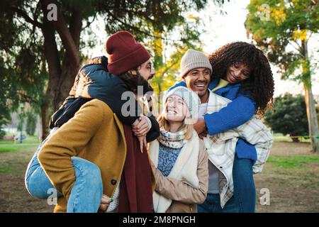Fünf Millennials-Freunde haben Spaß beim Huckepack im Park Outdoor. Eine Gruppe multiethnischer junger Menschen, die lächeln und ihre Freundinnen tragen, genießen einen Wochenendausflug. Lifestyle-Konzept. Hochwertiges Foto Stockfoto