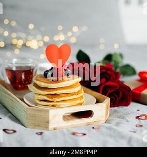 Zusammensetzung zum Valentinstag mit Frühstück, Geschenkbox und Rosen. Stapel köstlicher Pfannkuchen mit Beeren und eine Tasse heißer Mongungstee auf einem Holztablett. Stockfoto