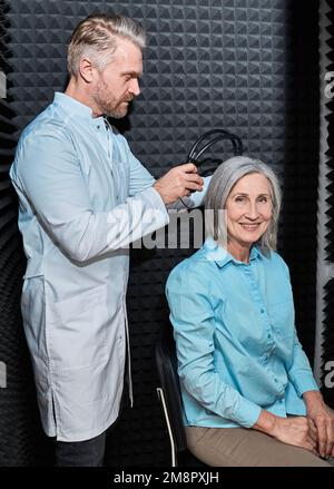 Audiometrie, Reintonaudiogramm-Test. Audiologe, der eine Höruntersuchung bei einer erwachsenen Frau in einer schalldichten Audiometerkabine mit Kopfhörer und Audiometer durchführt Stockfoto