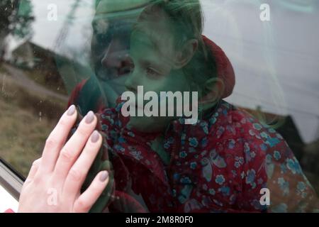 Porträt eines kleinen Mädchens, das im Auto sitzt und Auf Wiedersehen zu ihrer Mutter sagt. Flüchtlinge, Kriegskrise, humanitäres Katastrophenkonzept. Stockfoto