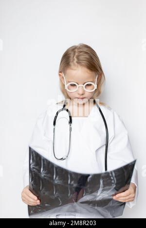 Kaukasisches Kind in Arztuniform mit Brille und Stethoskop, aufmerksam und ernst. Ein Kind, das sich einen Röntgenfilm ansieht Stockfoto