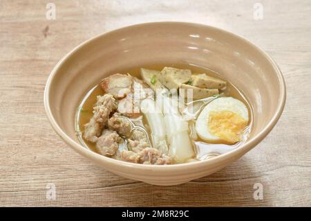 Große Reisnudeln, Wurstscheiben mit Kotelett und knusprigem Schweinefleisch, das weich gekochtes Ei in Suppe auf Schüssel serviert Stockfoto