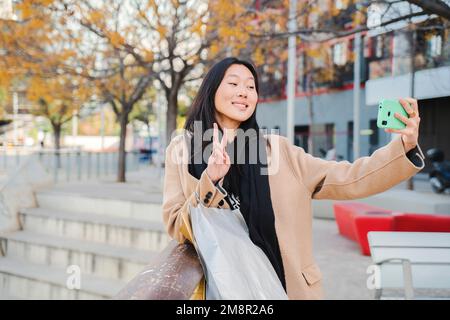 Fröhliche junge chinesin oder Asiatin, die lächelt und ein Selfie-Porträt macht, das Friedenszeichen auf dem Handy macht, nachdem sie eingekauft und über die sozialen Medien mit einer Smartphone-App geteilt hat. Hochwertiges Foto Stockfoto
