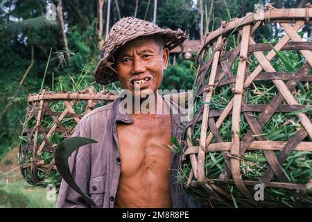 Horizontales Porträt südasiatischer balinesischer Senioren mit traditioneller kegelförmiger Mütze. Älterer Mann mit schiefen Zähnen, der in die Kamera schaut Stockfoto