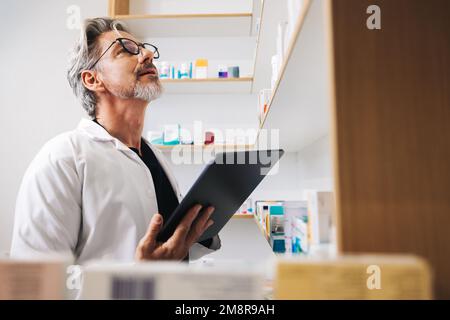 Apotheker auf der Suche nach verschreibungspflichtigen Medikamenten in einem Apothekenregal. Senior-Mann, der ein digitales Tablet benutzt, um Online-Rezepte in einer Apotheke auszufüllen. Stockfoto
