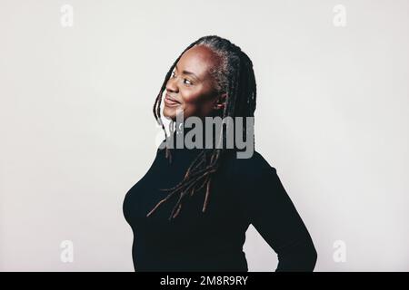 Eine nachdenkliche Frau mit Dreadlocks, die mit einem fröhlichen Lächeln in einem Studio wegsieht. Glückliche reife Frau, die sich Gedanken macht, während sie gegen einen grauen Hintern steht Stockfoto