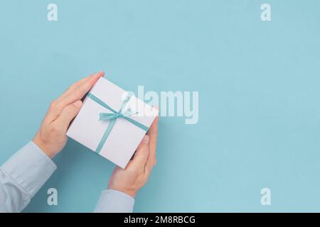 Hände halten Geschenkbox mit blauer Schleife mit Kopierplatz in Bastelpapier auf blauem Hintergrund. Draufsicht, flach liegend Stockfoto