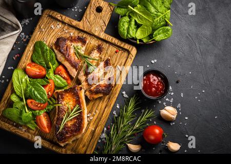 Schweinefleisch Steaks mit frischem Salat serviert auf Holzbrett. Draufsicht auf schwarzem Tisch. Stockfoto