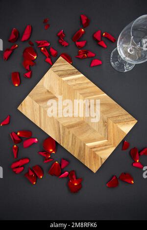 Holzbrett zum Kochen auf dunkelschwarzem Hintergrund mit einem Glas Wein und roten Rosenblättern. Ansicht von oben. Stockfoto