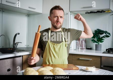 Gutaussehender Bäcker zu Hause, der am Tisch sitzt, mit einem Drehstift. Porträt eines massiven Kaukasier-Bäckers in grüner Schürzenuniform, der auf die Kamera schaut und Bizepsmuskeln zeigt. Hochwertiges Foto Stockfoto