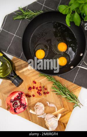 Schwarze Pfanne mit rohen Eiern auf einem dunklen karierten Handtuch neben einem hölzernen Kochbrett mit rotem Granatapfel und Knoblauch. Speisekonzept. Ansicht von bis Stockfoto