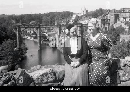 1950er, historisch, zwei gut gekleidete, reife Damen, die nebeneinander an der alten Burgmauer in Knaresborough, North Yorkshire, England, Großbritannien sitzen. Die Ruinen der königlichen Burg blicken auf den Fluss Nidd. Eine Brücke über den Fluss und die Stadt Knaresborough ist im Hintergrund zu sehen. Stockfoto