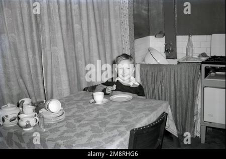 1950er. Historisch, in einer Küche, ein kleiner Junge mit einem großen Lächeln im Gesicht, sitzt an einem überdachten Tisch und trinkt Tee, ein Sandwich und ein Getränk, England, Großbritannien. Emaille-Schüssel im Waschbecken mit einem frei stehenden Gaskocher „New Home“ der Epoche daneben. Teetassen und kleine Teller auf dem Tisch. Stockfoto