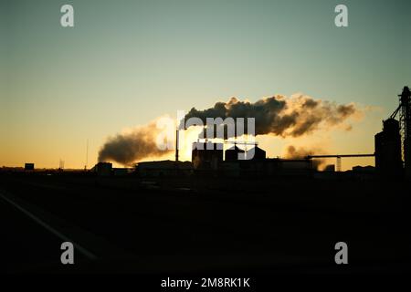 Eine Silhouettenaufnahme einer Fabrik, die Rauch aus ihren Schornsteinen auf dem Land emittiert Stockfoto