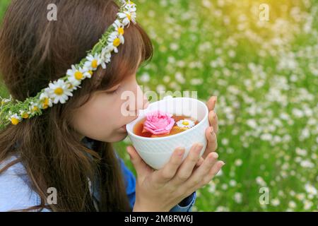 Brünett gesunde Frau, die duftenden Kräutertee trinkt. Selektiver Fokus. Ein Teenager im Blumenkranz, der einen weißen Becher mit Kamillentgiftungsgetränk hält Stockfoto