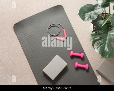 Stylische, graue und pinke, flache Heimfitness-Lay. Draufsicht auf graue Sportmatte, Yoga-Block, Springseil und rosa Hanteln auf neutralem Teppichhintergrund, Monstera-Pflanze. Set für Pilates, Fitness, Yoga Stockfoto