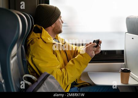 Ein bärtiger, lässiger Mann in gelber Jacke und Hut, der während der Fahrt mit dem Zug ein Smartphone benutzt, im Zug sitzt und aus dem Fenster schaut. Stockfoto