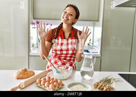 Junge, wunderschöne hispanische Frau, die selbstbewusst lächelt und in der Küche Hände mit Mehl zeigt Stockfoto