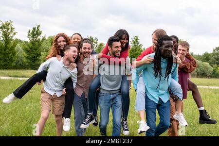 Eine Gruppe von besten Freunden und jungen Paaren, die Spaß im Park haben und Huckepackrennen machen, eine internationale Gruppe junger glücklicher Schüler beim öffentlichen Park Playi Stockfoto