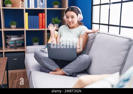Hispanische junge Frau, die zu Hause ein Notebook benutzt und Kopfhörer trägt, ohne Verstand und verwirrter Ausdruck, während Arme und Hände angehoben sind. Zweifelhaftes Konzept. Stockfoto