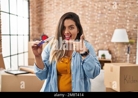 Junge hispanische Frau, die den Schwangerschaftstest in der neuen Wohnung hält, lächelt glücklich mit Hand und Finger Stockfoto