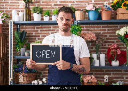 Junger lateinamerikanischer Mann, der beim Floristen arbeitet und das Schild offen hält, ist skeptisch und nervös, die Stirnrunzeln sind wegen des Problems aufgebracht. Negative Person. Stockfoto