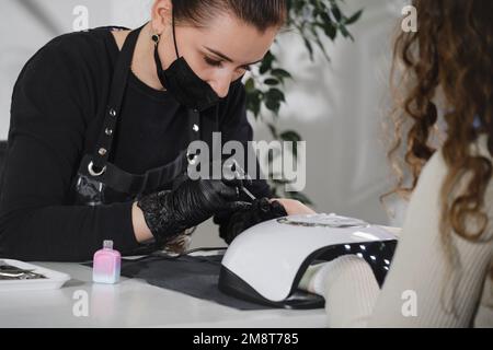 Maniküre-Salon. Professionelle Maniküre mit Schutzmaske, die sich an Frauen im modernen Schönheitssalon richtet. Konzept der Schönheitsindustrie. Lampe für Stockfoto