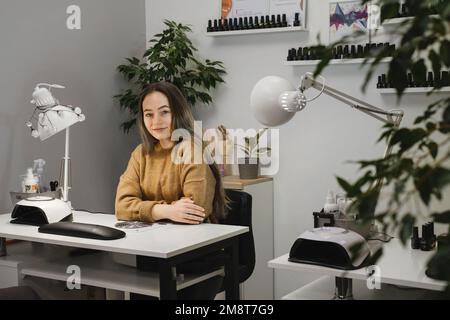 Nagelmeister. Porträt einer professionellen jungen Brünette, die am modernen Nagelsalon sitzt und lächelt, während sie auf den Kunden wartet. Beruf Stockfoto