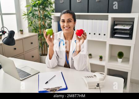 Junge hispanische Frau, die in einer Diätistischen Klinik arbeitet, hält einen grünen Apfel und lächelt mit einem fröhlichen und kühlen Lächeln im Gesicht. Zähne zeigen. Stockfoto