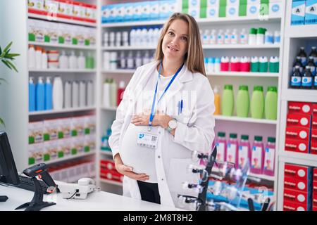 Junge schwangere Apothekerin, die selbstbewusst lächelt und den Bauch in der Apotheke berührt Stockfoto