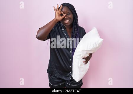 Junge afrikanische Frau mit Pijama-Umarmungskissen, die eine gute Geste mit lächelnder Hand macht, mit einem Auge, das durch die Finger blickt, mit fröhlichem Gesicht. Stockfoto