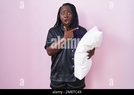 Junge afrikanische Frau mit Pijama-Umarmungskissen überraschend mit dem Finger zur Seite zeigen, offener Mund erstaunlicher Ausdruck. Stockfoto