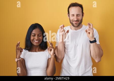 Ein gemischtrassiges Paar, das über gelbem Hintergrund steht, mit dem Finger lächelt, mit Hoffnung und geschlossenen Augen. Glück und abergläubisches Konzept. Stockfoto