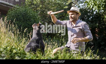 Bärtiger Tierbesitzer mit Strohhut, der im Sommer draußen mit seinem Hund spielt. Stockfoto