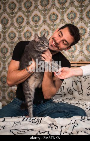 Der junge Mann spielt mit seiner Katze auf dem Bett. Der schöne Mann streichelt die flauschige graue Katze. Stockfoto