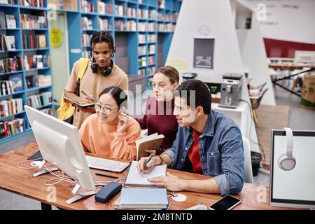 Vielfältige Gruppe von Studenten, die Computer zusammen in farbenfrohen Bibliotheksräumen nutzen und gerne lernen Stockfoto