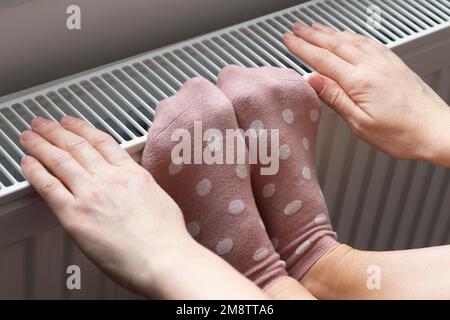 Eine Frau wärmt ihre Füße und Hände in Socken auf einem Heizkörper. Heizelemente. Niedrige Temperatur und Kälte im Haus. Foto-Nahaufnahme Stockfoto